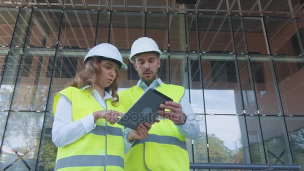 Man och kvinna ingenjörer har samtal och använda Tablet PC: N på byggarbetsplatsen. Glasbyggnad på bakgrund. — Stockvideo