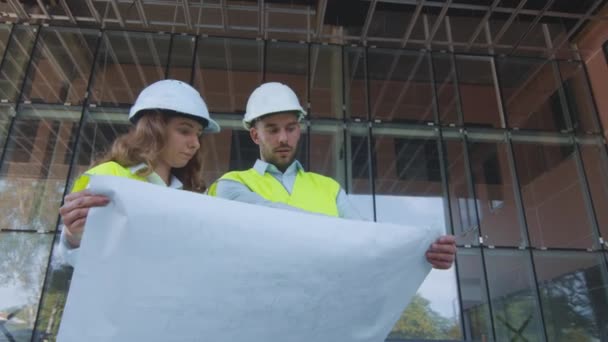 Man en vrouw ingenieurs hebben gesprek en op zoek naar grote blauwdruk. Glazen gebouw op achtergrond. — Stockvideo