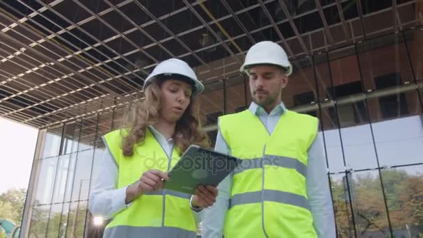 Équipe de deux ingénieurs marchant, parlant et utilisant un ordinateur tablette. Bâtiment en verre ou gratte-ciel en construction sur fond . — Video