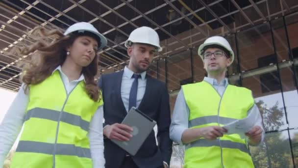 Team av två ingenjörer och affärsman i hård hatt promenader, prata och använda Tablet PC: N. Glasbyggnad eller skyskrapa under konstruktion på bakgrunden. — Stockvideo