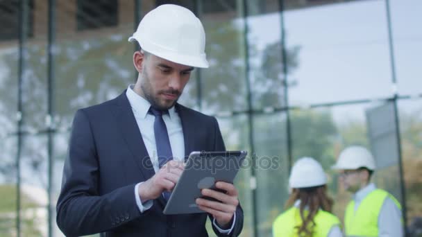 Empresário em Hard Hat Caminhando, Falando, e Usando Tablet Computer. Edifício de vidro ou arranha-céu em construção no fundo . — Vídeo de Stock