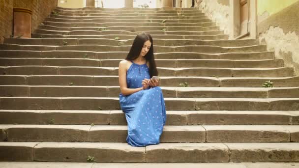 Atractiva joven morena en vestido de verano ligero sentada en las escaleras de la ciudad europea. Ella está mirando su teléfono móvil y usarlo . — Vídeo de stock