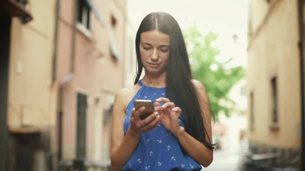 Attraktiv ung brunett kvinna i ljusa sommarklänning på gatorna i Europeiska stad. Hon tittar på sin mobiltelefon och använda den. — Stockvideo
