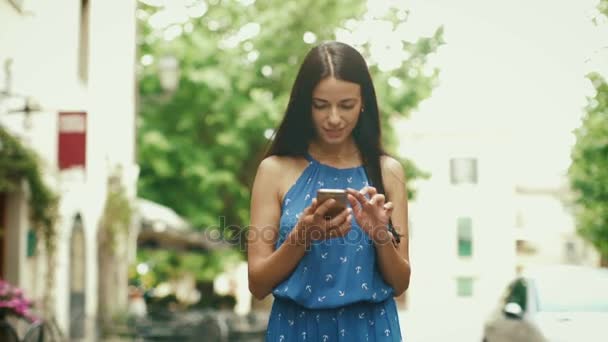 Jovem morena atraente em luz vestido de verão andando nas ruas da cidade europeia. Ela está olhando para seu telefone celular e usá-lo . — Vídeo de Stock