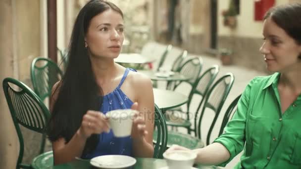Две привлекательные молодые женщины в светлых летних платьях расслабляются и беседуют в кофейне на улице . — стоковое видео