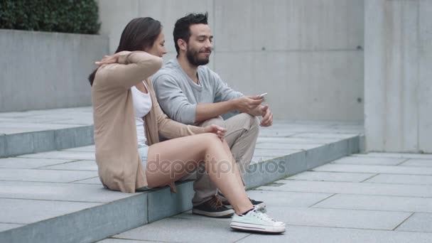 Молодой счастливый улыбающийся мужчина и женщина сидят на ступеньках на улице и общаются . — стоковое видео