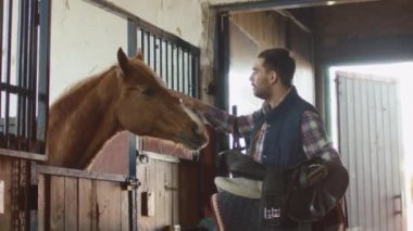 Adam bir at ahırda bir eyer tutarken okşayarak.