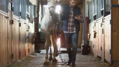Adam ahırda bir cep telefonu kullanırken beyaz bir at yürüyor.