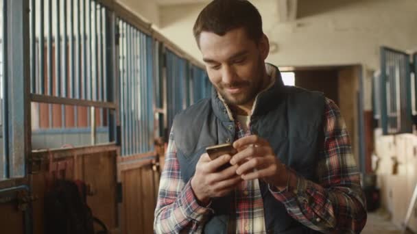 Feliz hombre sonriente está utilizando un teléfono inteligente junto a un caballo en un establo . — Vídeos de Stock