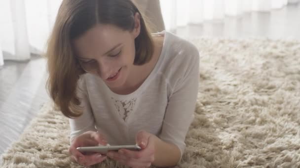 Femme attrayante posée sur le sol dans le salon et utilisant le téléphone portable pour le divertissement . — Video