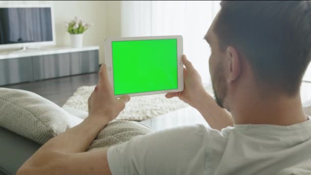 L'uomo è sdraiato sul divano a casa e guardare sul tablet con schermo verde in modalità paesaggio . — Video Stock