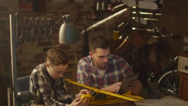 Vader en zoon zijn een speelgoed vliegtuig modelleren terwijl het controleren van de instructie op een tablet-pc in een garage bij u thuis. — Stockvideo