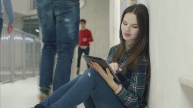 Genç çekici kız öğrencinin üniversite koridorda otururken bir tablet bilgisayar kullanarak portresi.