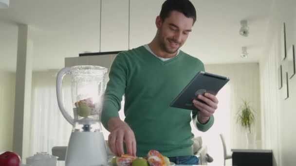 Bonito homem está preparando um smoothie ao usar um computador tablet na cozinha . — Vídeo de Stock