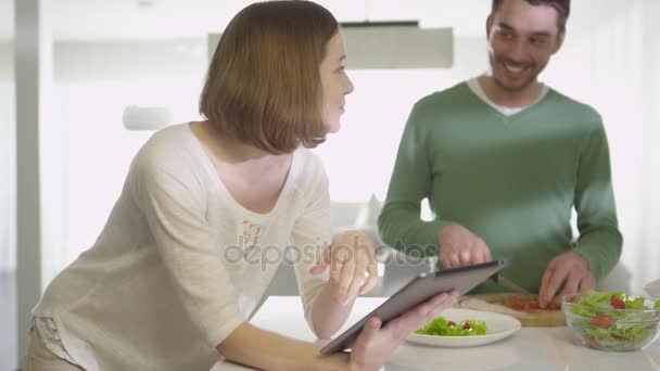 Casal feliz está preparando comida saudável na cozinha em casa enquanto verifica um computador tablet . — Vídeo de Stock