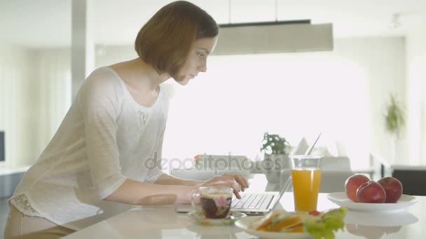 Jovem mulher está usando um computador portátil de manhã na área de cozinha em casa . — Vídeo de Stock