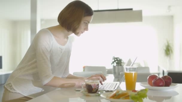 Jonge vrouw is een laptopcomputer het gebruik in de ochtend bij huis keuken. — Stockvideo