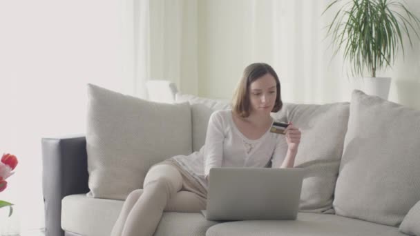 Jovem mulher está sentada no sofá em casa e compras on-line usando cartão de crédito . — Vídeo de Stock