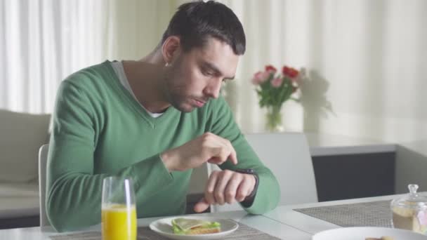 El hombre está revisando su reloj inteligente mientras almuerza y toma jugo de naranja por la mañana. — Vídeo de stock