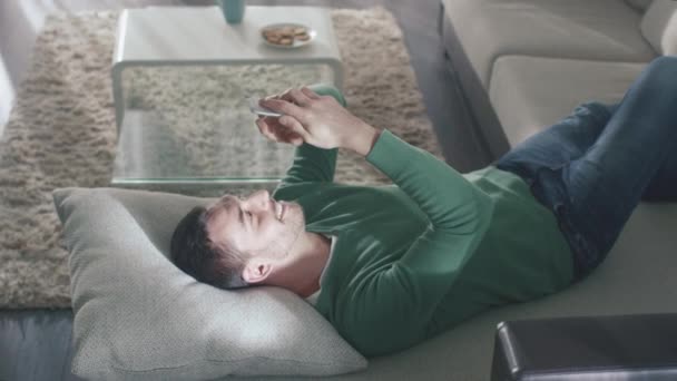 Homem está deitado no sofá em casa enquanto vestindo jeans e casaco verde e usando smartphone . — Vídeo de Stock