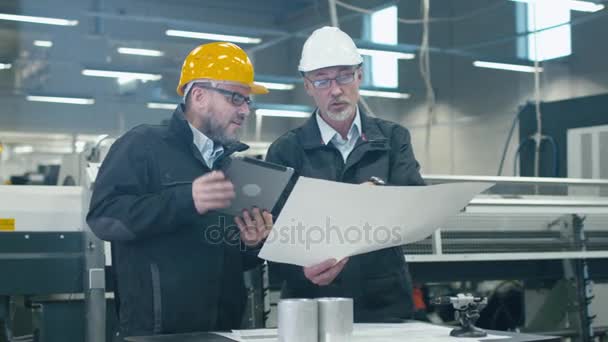 Dos ingenieros discuten un plan mientras verifican la información en una tableta en una fábrica . — Vídeo de stock