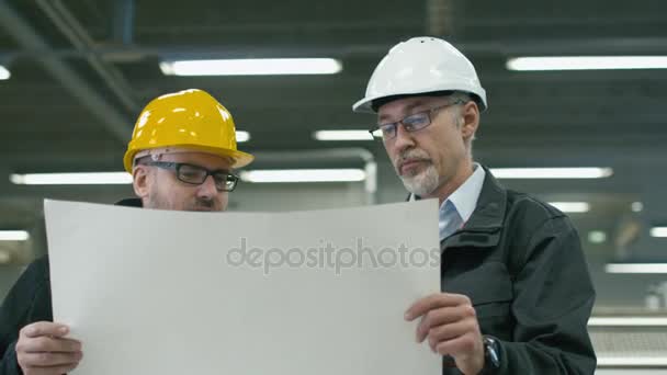 Twee ingenieurs in hardhats bespreken een blauwdruk terwijl je in een fabriek. — Stockvideo