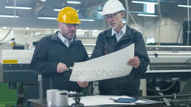 Two engineers in hardhats discuss a blueprint while standing in a factory. — Stock Video