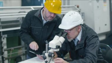 Kıdemli mühendis bir fabrikada mikroskop altında bir detay inceliyor. İkinci çalışan veri bir tablet bilgisayar koyar..