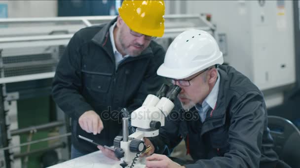 Старший инженер осматривает детали под микроскопом на заводе. Второй сотрудник помещает данные в планшетный компьютер . — стоковое видео