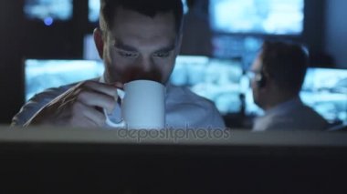 Mutlu teknik destek uzmanı karanlık bir bilgisayarda çalışırken kahve içiyor Oda izleme göstermek perde ile dolu.