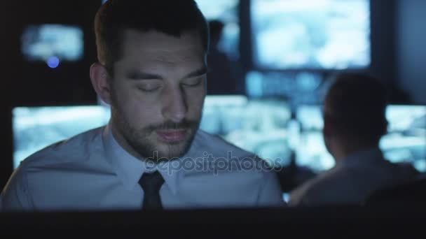Veiligheidsbeambte is praten over de telefoon terwijl het werken op een computer in een donkere controle kamer gevuld met schermen. — Stockvideo