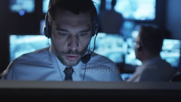 Spécialiste du support technique parle sur un casque tout en travaillant sur un ordinateur dans une salle de surveillance sombre remplie d'écrans d'affichage .