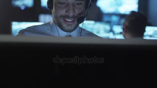 El especialista en soporte técnico está hablando con auriculares mientras trabaja en una computadora en una sala de monitoreo oscura llena de pantallas . — Vídeos de Stock