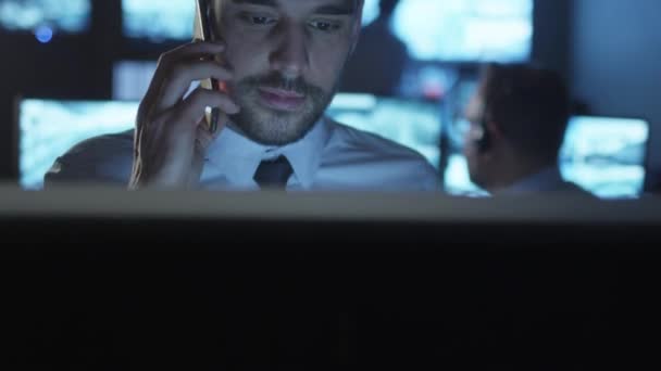Oficial de segurança está falando ao telefone enquanto trabalha em um computador em uma sala de monitoramento escuro cheio de telas de exibição . — Vídeo de Stock