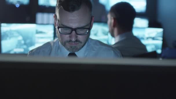 Feliz empleado de oficina en camisa blanca está sentado junto a una computadora, mirando a la cámara y sonriendo en una oscura sala de monitoreo llena de pantallas de visualización . — Vídeos de Stock