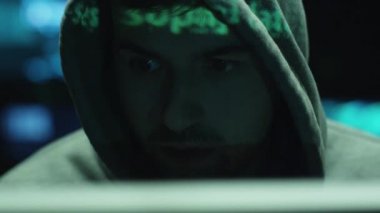 Yeşil kod karakterleri karanlık bir ofiste yüzünü yansıtırken, erkek hacker bilgisayar üzerinde çalışıyor..