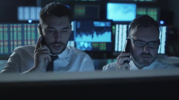Dva makléři jsou aktivně mluví o telefonech, zatímco pracují na počítačích v tmavé kanceláři plné obrazovky. — Stock video