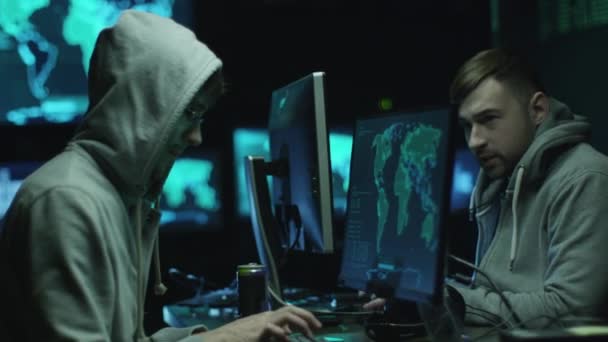 Dois hackers com capuz trabalham em computadores com mapas e dados em telas de exibição em uma sala de escritório escura . — Vídeo de Stock