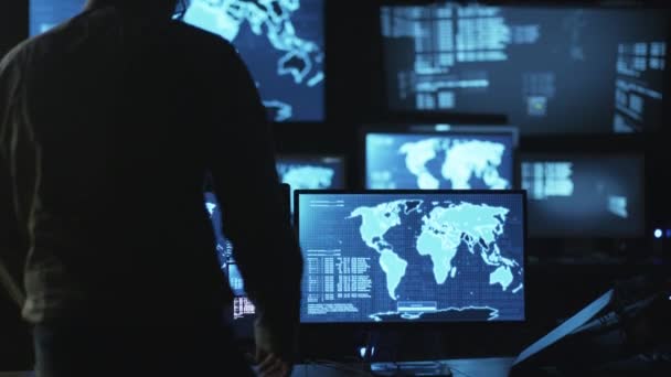 Employé informatique masculin vient à la salle de surveillance pour travailler sur ordinateur avec des cartes et des données affichées dans une salle de bureau sombre .