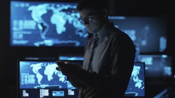 Mann mit Brille benutzt Smartphone in dunklem Überwachungsraum voller Bildschirme. — Stockvideo