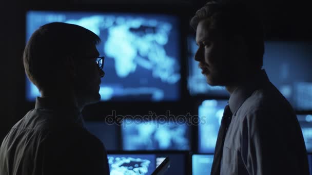 Twee mannelijke werknemers ondervindt een gesprek in een donkere controle kamer gevuld met schermen. — Stockvideo