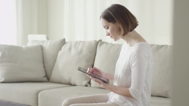 Bella giovane donna sta usando un computer tablet mentre seduto su un divano a casa . — Video Stock