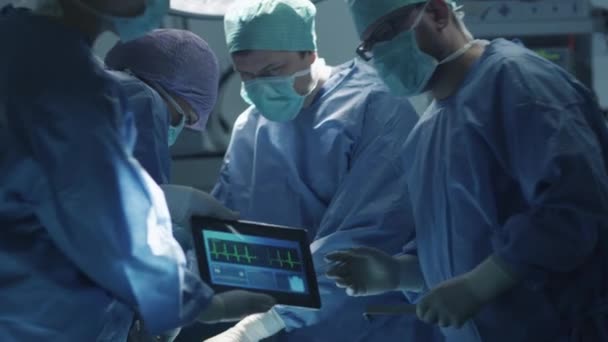 Медицинская команда, выполняющая хирургическую операцию в современной операционной. Использование планшета для получения дополнительной информации . — стоковое видео