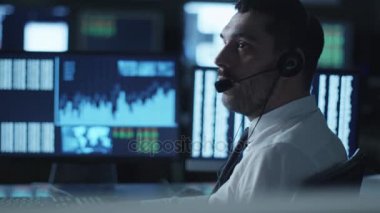 Karanlık bir ofiste bir bilgisayarda çalışma göstermek perde ile dolu iken borsacı bir kulaklık üzerinde konuşuyor.