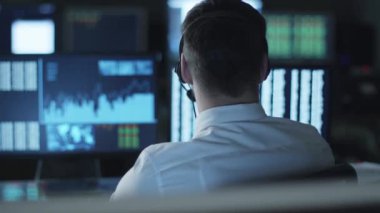 Mutlu başarılı borsacı bir kulaklık ile bir sandalyeye dönme ve göstermek perde ile karanlık bir ofiste gülümseyen.