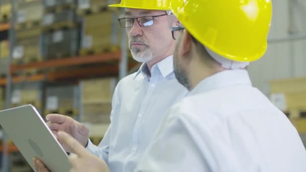 Dos empleados en el almacén del centro logístico están discutiendo el trabajo mientras sostienen una computadora portátil . — Vídeos de Stock