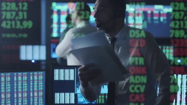 Makler giełdowy w białej koszuli jest rozmawia przez telefon podczas pracy w ciemnym pokoju monitorowania z ekranów. — Wideo stockowe