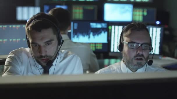 Два биржевых брокера разговаривают на гарнитуре, работая за компьютером в темном офисе, заполненном дисплейными экранами . — стоковое видео