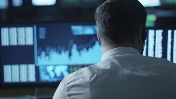Stockbroker розмовляє на гарнітурі під час роботи на комп'ютері в темному офісі, наповненому екранами дисплеїв . — стокове відео