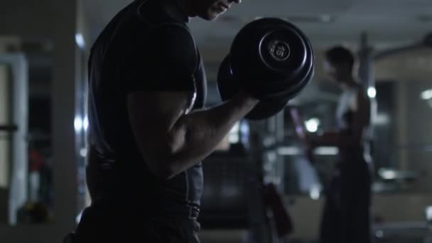 Handsome fit sporty man does dumbbell curl exercises in dark gym. — Stock Video